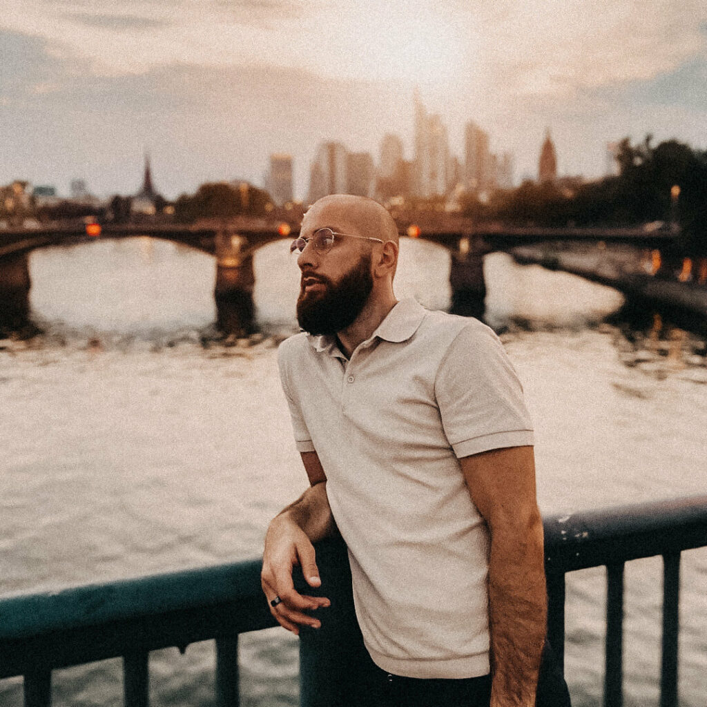 Isa Ulubaev lehnt sich an ein Brückengeländer an und schaut nachdenklich nach links entlang der Brücke. Im Hintergrund ist eine Skyline zu sehen