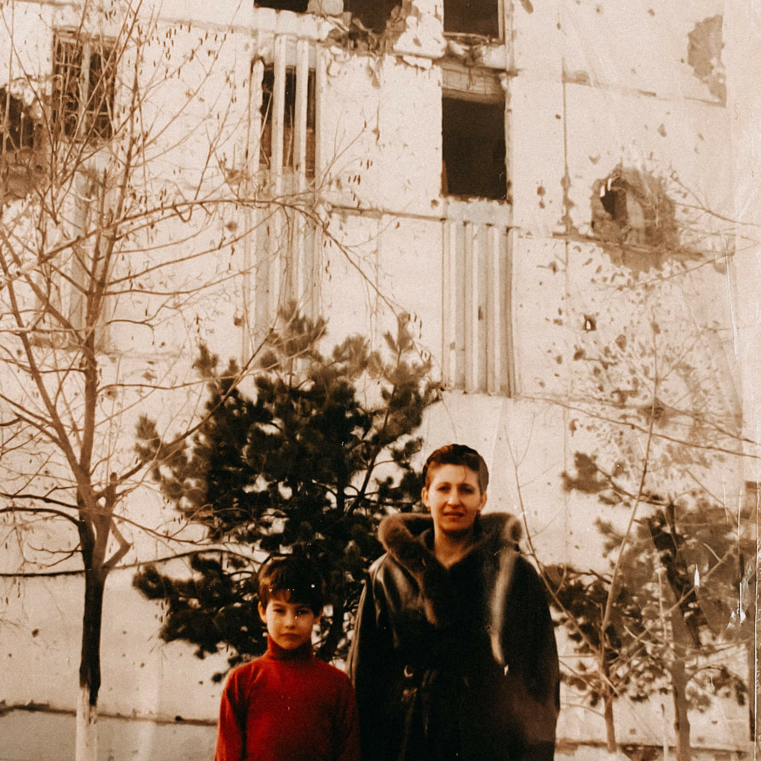 Isa Ulubaev als Kind mit seiner Mutter die einen Mantel trägt. Im Hintergrund ist ein zerstörtes Gebäude zu sehen